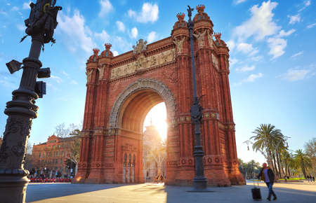 Sunrise at Triumphal Arch in Barcelona, Catalonia, Spain. Arc de Triomf at boulevard street. Alley with tropical palm trees. Early morning landscape with shadows and blue sky with clouds. Famous landmark.の写真素材
