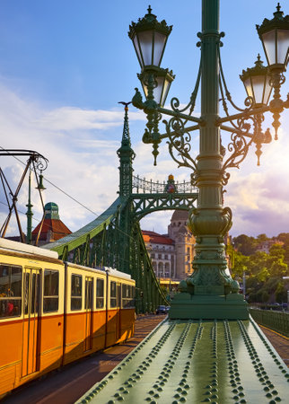 Budapest, Hungary. Old tram on Freedom bridge with sunlight in sunset sky, clouds and pole with street lamps, evening urban landscape. Travel Europe.の写真素材