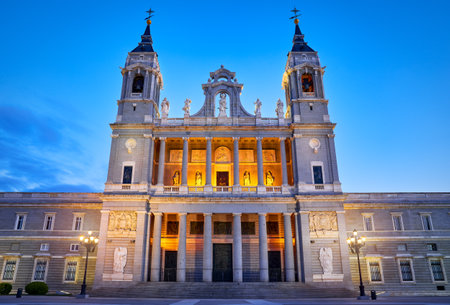 Madrid, Spain. Front view of Cathedral Santa Maria la Real de la Almudena at Plaza de la Armeria. Famous landmark with sunset sun, flowers and green bush. Street lamps with illumination and blue sky.の写真素材