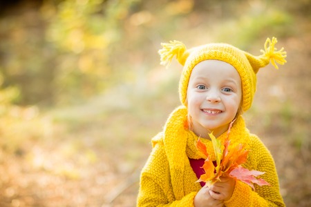 Cute little girls with big bue eyes playing on beautiful autumn day. Happy children having fun in autumn park. The girl is dressed in yellow coat and a hat. She holds a bouquet of yellow leaves in her handの写真素材