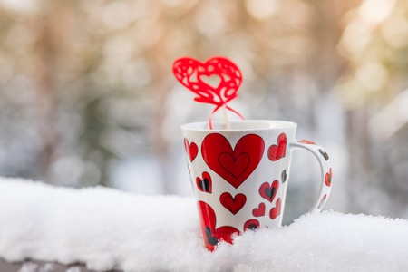 Love at coffe time, christmas concept, mug and red heart decoration on a snow balcony.Cup with heart and a hot drink wrapped in a scarf in the snow.Valentines day conceptの写真素材