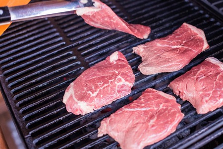 Man placing raw meat on to a hot barbecue to grill using a pair of wooden tongs, close up view over a dark background with copy space.raw meat, grill,bbq season. summer foodの写真素材