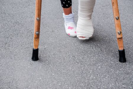 Young girl in orthopedic cast on crutches walking on the street near the road. Child with a broken leg on crutches, ankle injury. Bone fracture and ankle fracture in childrenの写真素材