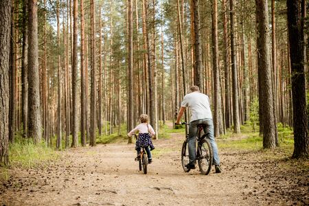 back view of father and daughter riding bicycles around forest.Young family in summer cycling in park.The theme family sports outdoor recreation.Adventure leisure concept.の写真素材
