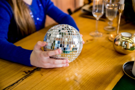 Mirrored shiny silver disco ball in kids hands on christmas festive table.girl holding silver disco ball, table decoration . Concept party, holiday. Copy spaceの写真素材