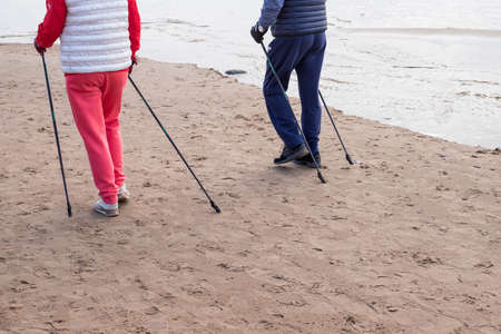 senior nordic pole walker enjoying nice autumn day outdoors, near sea. active elderly man and woman wearing sport clothes having walk by lake using specially designed poles. Selective focusの写真素材