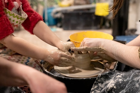 A close-up of a woman potter teaches a child how to properly mold a bowl of brown clay on a potters wheel in a bright workshop. Training of modeling on a potters wheel.child sculpts with clayの写真素材
