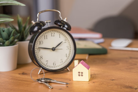 Model house on wooden table, keys, red alarm clock on working place. Real Estate Concept.
