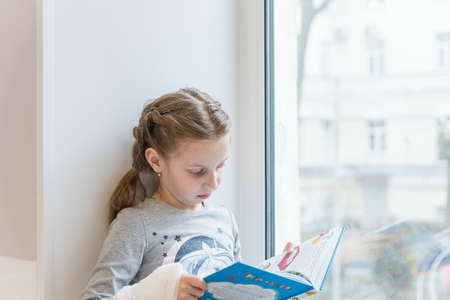 Cute little girl reading book on windowsill.Portrait of beautiful little European girl having concentrated look, resting on windowsill.studying indoor concept, clever schooler, daytime.の写真素材