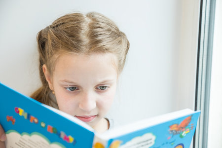 Cute little girl reading book on windowsill.Portrait of beautiful little European girl having concentrated look, resting on windowsill.studying indoor concept, clever schooler, daytime.の写真素材