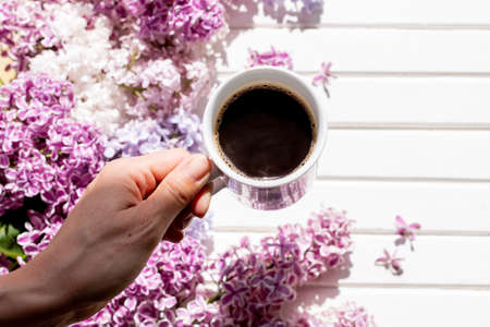 coffee mug in hand.Hands of young woman drinking coffee on white wooden table with flowers, gift. Woman hand hold cup of black coffee, spring flowers, lilac bush. Spring mood. Beautiful breakfastの写真素材