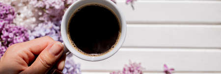 coffee mug in hand.Hands of young woman drinking coffee on white wooden table with flowers, gift. Woman hand hold cup of black coffee, spring flowers, lilac bush. Spring mood. Beautiful breakfastの写真素材