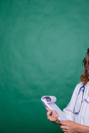 Doctor filling medical report for her patient.Doctor with stethoscope in white coat holding clipboard, writing medical record diagnosis, isolated on green background, close up.copy spaceの写真素材
