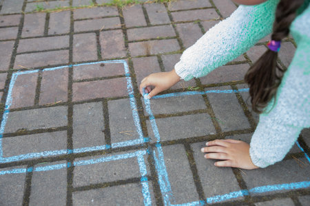 girl draws a hopscotch with chalk on the pavement.Child playing the game outside. Kid wearing...