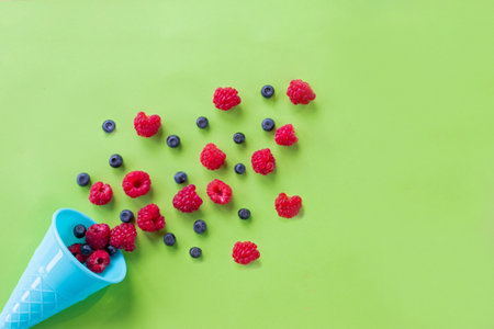 Waffle sweet ice cream cone with raspberries over white wooden background, top view. Flat lay.Creative food concept with colorful cone on pastel background. Mix of summer berriesの写真素材