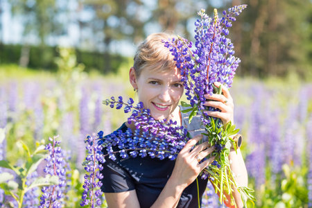 Portrait of young woman holding bouquet of lupin flowers walking in summer meadow. Stylish girl is enjoying sunny weather. blonde woman with bouquet of flowers.Lupine field. Summer vacation.の写真素材