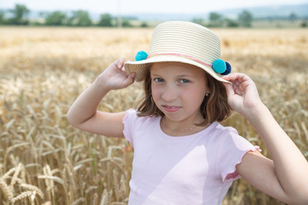 little blonde girl in a hat and a pink dress, standing on a wheat field and touching the ears of wheat with her hands. child enjoying nature wheat field.Happy momentsの写真素材