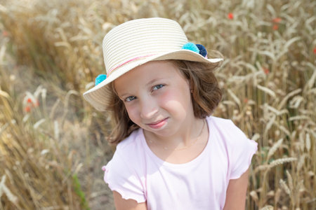 little blonde girl in a hat and a pink dress, standing on a wheat field and touching the ears of wheat with her hands. child enjoying nature wheat field.Happy momentsの写真素材