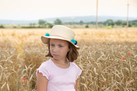 little blonde girl in a hat and a pink dress, standing on a wheat field and touching the ears of wheat with her hands. child enjoying nature wheat field.Happy momentsの写真素材