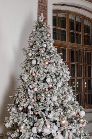 Artificial Christmas tree in a bright interior with silver, pink, bronze balls and snow on the branches. The concept of New year and Christmas. Luxury Christmas tree with glowing garland.の写真素材