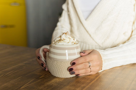 Morning coffee with whipped cream. winter composition. Hand with a cup of Cappuccino or latte coffee on christmas lights background.mug of hot cocoa in the hands of a girl with a dark red manicureの写真素材