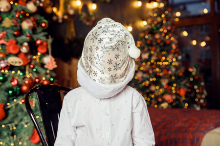 unrecognizable child in white shirt and santa hat stands on christmas tree background. Christmas celebration and fun.Faceless portrait. Boy with Christmas hat on his face.の写真素材