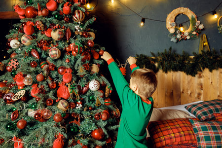 unrecognizable Child hanging a ball on illuminated Christmas tree.glowing lights. Boy in green pajamas decorating Christmas tree.Christmas good mood. Lifestyle, family and holiday.の写真素材