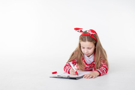 Merry Christmas and Happy Holidays. Happy child girl dreams of a gift and writes a letter to Santa Claus, lying on white background.Dreamy little girl in red xmas sweater and festive headbandの写真素材