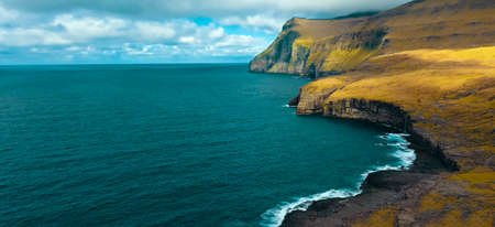 Faroe Islands, Flying around small grassy island with beautiful scenic mountains and cliffs overlooking the tranquil deep blue ocean. Stunning shot of islet in the picturesque Scandinavian countrysideの写真素材
