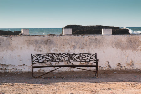 Empty iron designed bench on the banks of Atlantic ocean. Old white wall in the background, rocks...