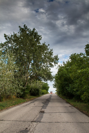 Country road among the trees of little forest. Red bus of public transport with the lights on on the horizon. Intense stormy clouds.の写真素材