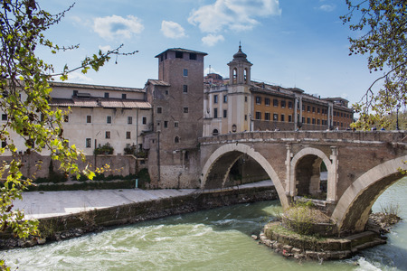 Green water of river Tiber, surrounding the Island with historical buildings and an old bridge. Cloudy blue sky.の写真素材