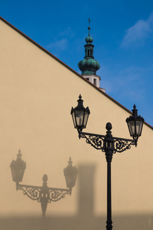 Traditional lantern with its shadow on the yellow diagonal wall. In the background tower of a church. Boskovice, Czech republic.の写真素材