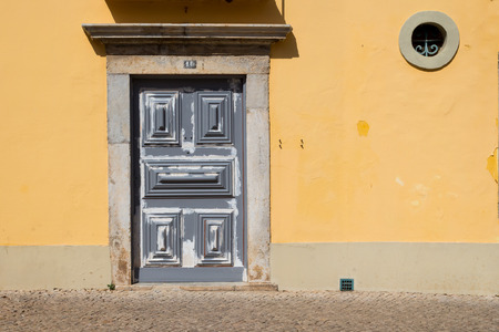Closed grey door of an old yellow house. Round small window. Street full of sunshine. Faro, Portugal.の写真素材