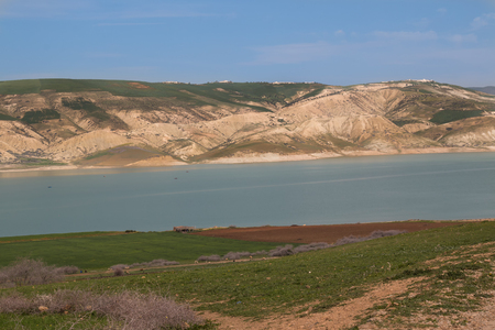 Calm water of lake Barrage Idriss in Morocco, close to Fez. Field in the foreground, mountain in the background. Blue cloudy sky.の写真素材