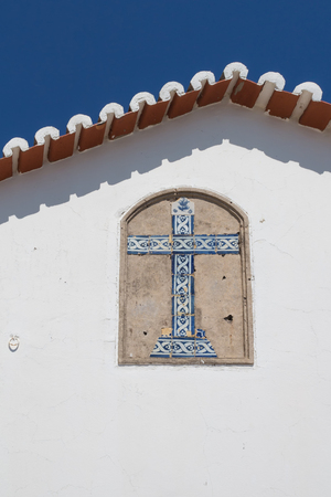 White side wall of a church of Nossa Senhora do Rocha with a cross made of traditional ceramics tiles - azulejos. Edge of the roof. Bright blue sky. Porches, Algarve, Portugal.の写真素材