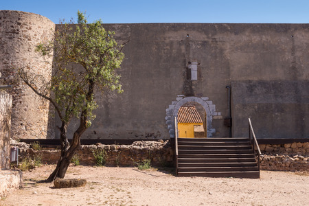 Square with an old tree and fortification stone wall with a gate with an arch. Wooden stairs. Bright blue sky. Castro Marim, Algarve, Portugal.のeditorial素材