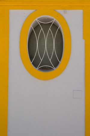 White wall of a house with oval window with a grid. Window lined by yellow painted frame. Next yellow lines. Vila Real de Santo Antonio, Algarve, Portugal.の写真素材