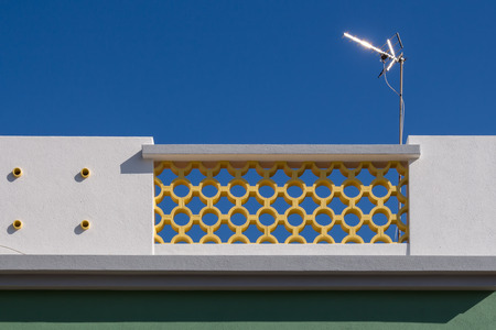 Fence of a roof terrace. Simple white facade and a yellow part with pefrorated decoration. Glossy new antenna. Clear blue sky. Alvor, Algarve, Portugal.の写真素材