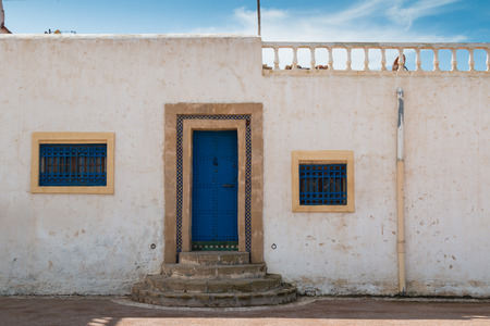 Street with a house with bright blue door and window grids and white color of the facade. Rounded stairs.  Terrace on the roof. Blue sky with white clouds. Street in Kasbah de Oudaias, Rabat, Morocco.の写真素材