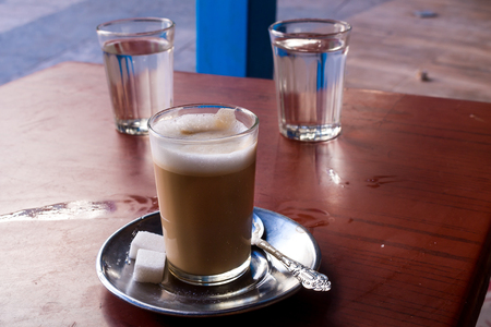 Traditional moroccan coffee with milk in a simple glass. Plate, ornate spoon and sugar cubes. Dirtly table on a terrace in the street. Safi, Morocco.の写真素材