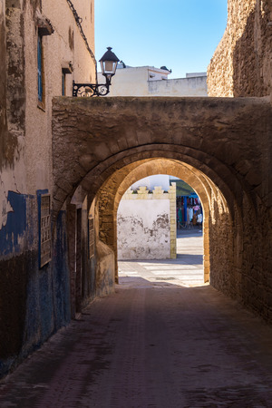 Old city with its typical narrow street, lined by houses and a wall on the other side. Undepass with an arch. Morning blue sky. Safi, Morocco.の写真素材