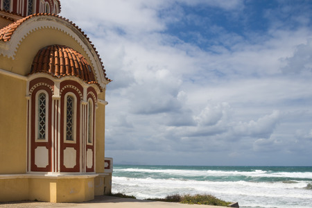 Coloroful small orthodox church of Ag. Fotini, located on the seaside. Foamy waves and horizon of the sea. Cloudy autumn sky. Rethymno, Crete, Greece.の写真素材