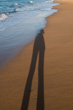 Warm yellow color of a clean sand of a beach. Small waves of the sea. Long woman shadow in the late afternoon sunlight. Heraklion, Crete, Greece.の写真素材