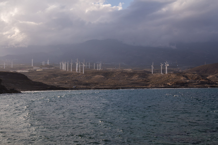 View from the coast on the Atlantic ocean waves. Wind electricity power stations, city and a mountain in the background. Cloudy sky. Punta de Abona, Tenerife, Canary Islands, Spain.の写真素材