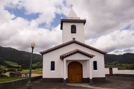 Church in a small village, surrounded by fields and mountains. White facade, tower with a cross. Intense cloudy sky. Lomba do Pomar, Sao Miguel, Azores Islands, Portugal.の写真素材