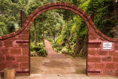 Stone gate with an arc, painted red, entrance to the botanic garden Ribeira do Guilherme. Fresh plants and trees lining the footpath. Nordeste, Sao Miguel, Azores Islands, Portugal.の写真素材