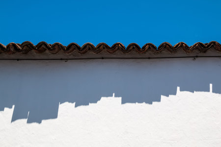 Wavy edge of a roof with its shadow on a white painted facade of a house. Bright blue sky. Ribeira...