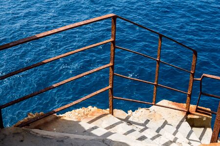 Stairs on a cliff at Neptuns Cave (Grotto di Nettuno). Rusted railing. Calm deep blue water of Mediterranean sea. Sardinia, Italy.の写真素材