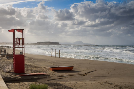 Sandy beach with a lifeguard tower and a red boat. No people in the autumn, during a rainy and stormy day. Waves of the sea and intense clouds. Heraklion, Crete, Greece.の写真素材